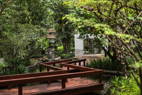 Wooden bridge crossing a landscaped water garden beneath mature tropical trees